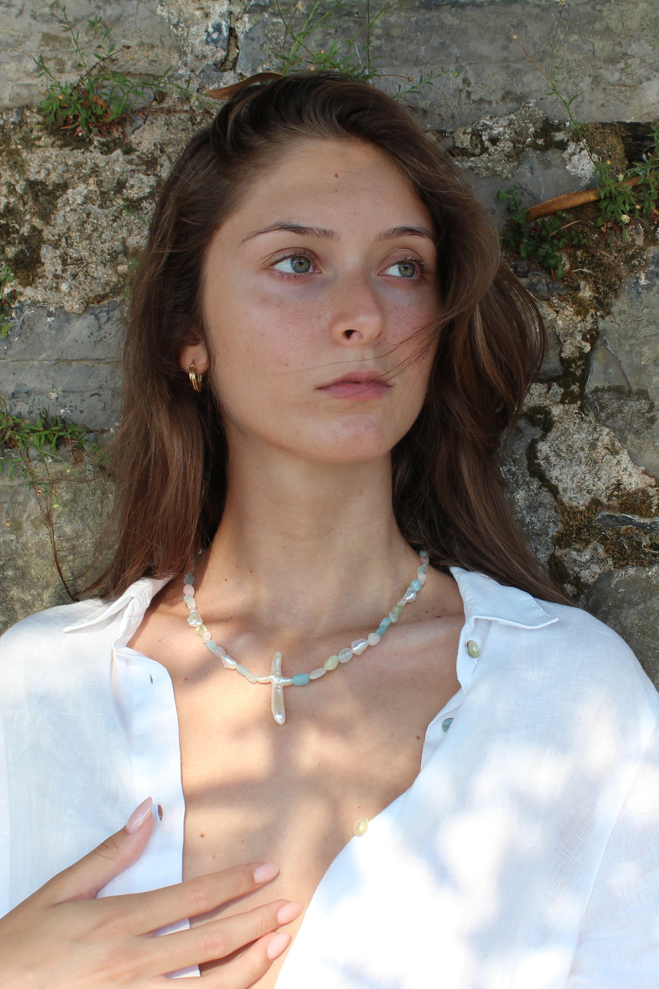 Woman in a white shirt standing against a stone wall wearing a gemstone necklace with a pearl cross