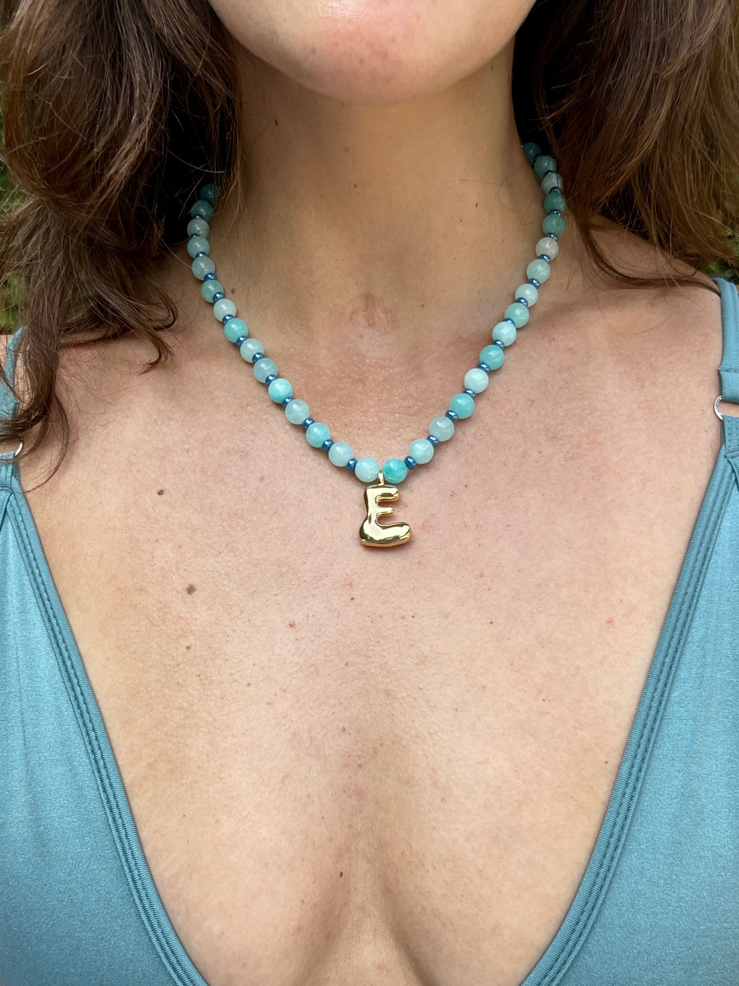 Person wearing a blue top and turquoise beaded necklace with a initial pendant.